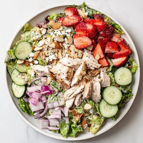 Strawberry Poppy Seed Chicken Salad served in a white bowl, garnished with toasted almonds, crumbled feta, and sliced cucumbers on a rustic wooden table.